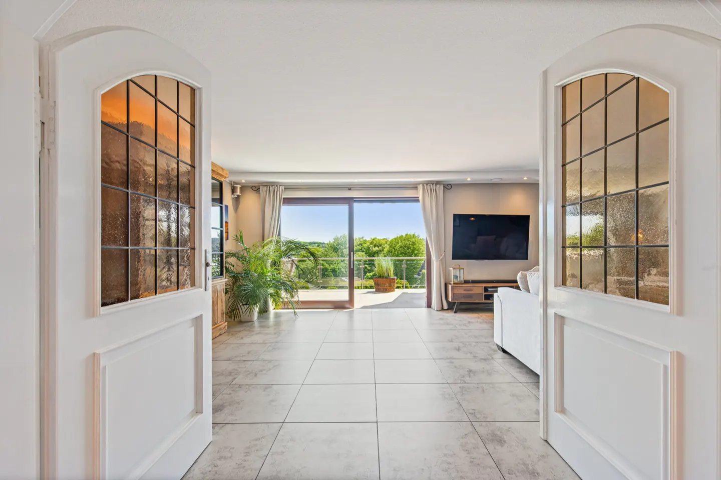 Open white doors reveal a bright living room with a balcony view. Gray tile floors, a TV, and a white sofa are visible.