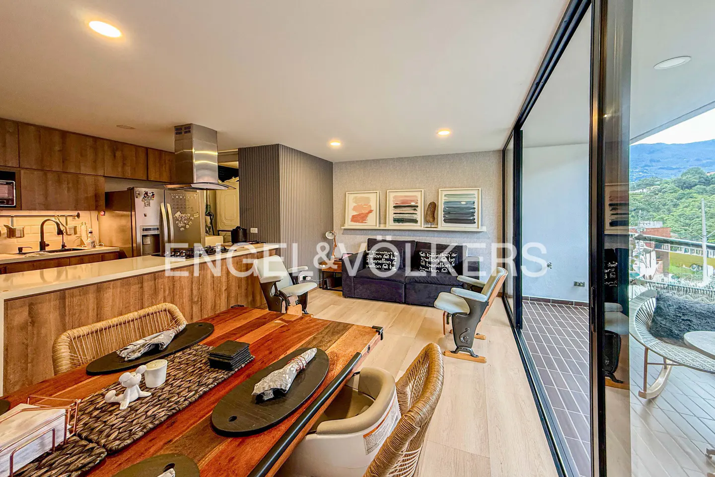 Modern open-plan living area with wood kitchen, dining table, dark sofa, and balcony with mountain views.