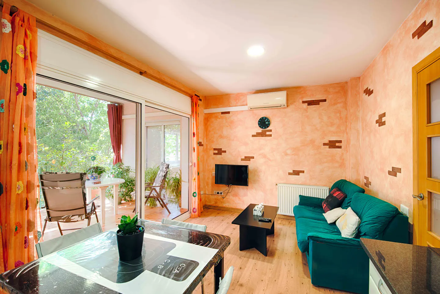 Bright living room with orange walls, a green sofa, and a balcony with outdoor seating. A black coffee table sits in front of the sofa.