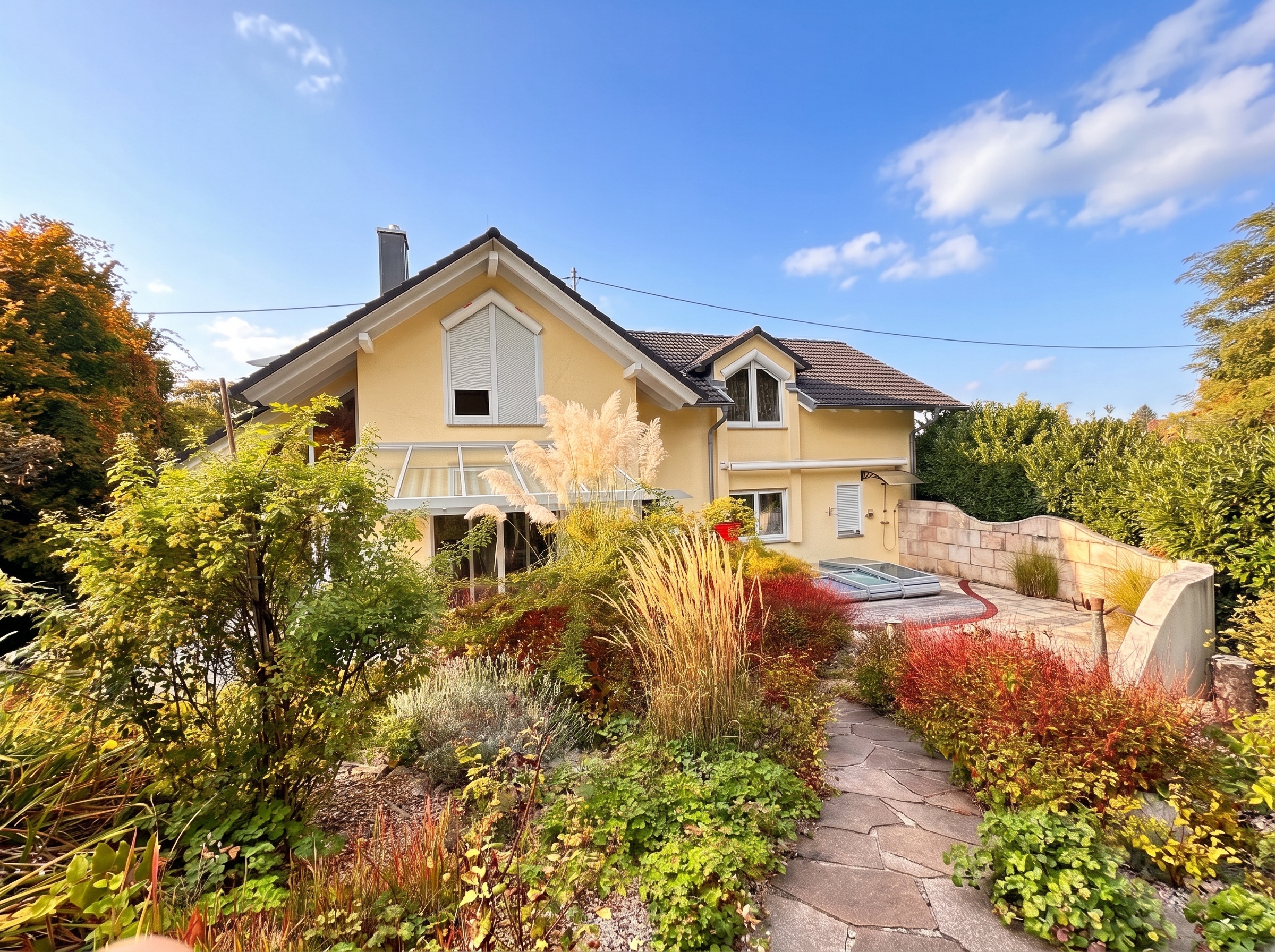 A yellow two-story house with a stone patio and a flagstone path surrounded by colorful autumn foliage and tall grasses. [AI-generated text]