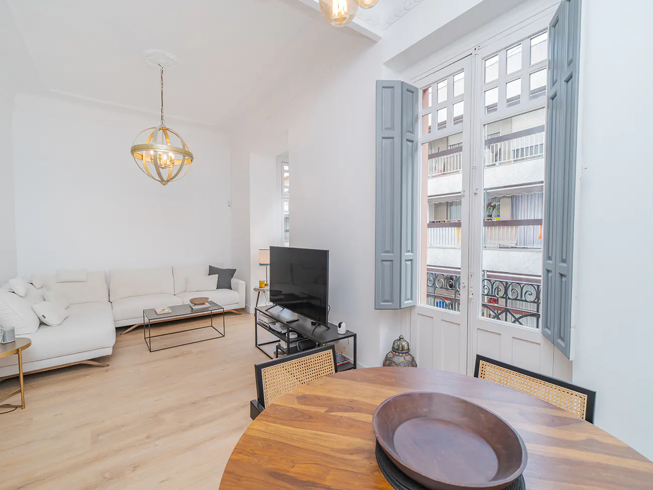Bright living room with white walls, light wood floors, and a large window with gray shutters. A round wood table sits in the foreground.