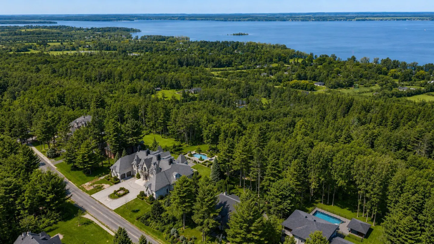 Aerial view of a large gray house with a pool, surrounded by green trees and a blue lake in the background.