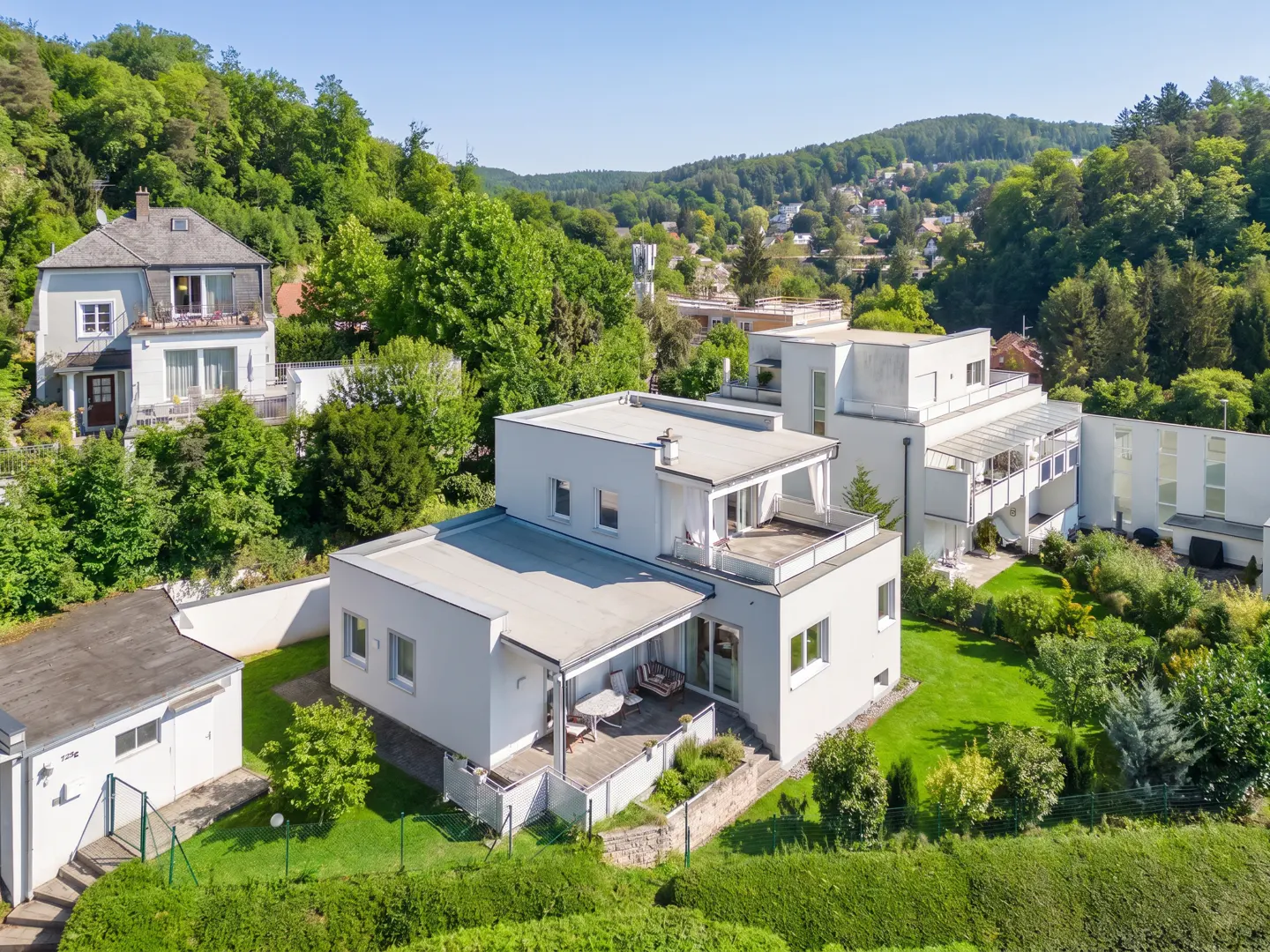 Exterior of a modern, multi-story white house with a flat roof, surrounded by green trees and hills under a blue sky.