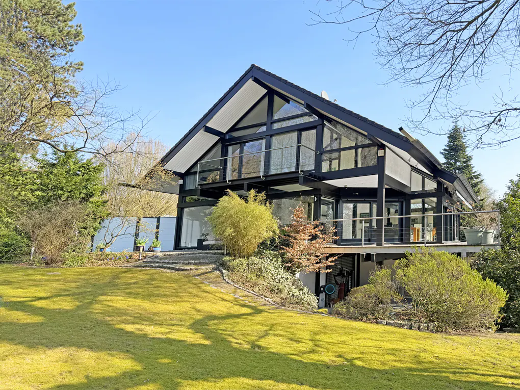 Two-story modern house with black trim and large windows, set on a green lawn with trees and shrubs.