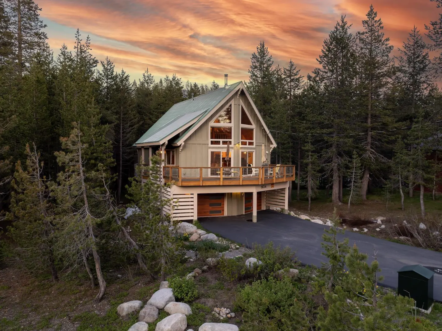 A two-story A-frame cabin with a green roof and wooden deck, nestled in a pine forest at sunset.