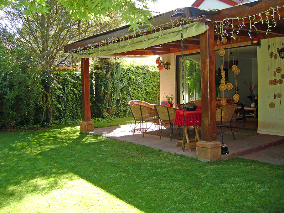 A backyard patio with a wooden pergola, string lights, and wicker furniture. A green lawn and hedge surround the patio.