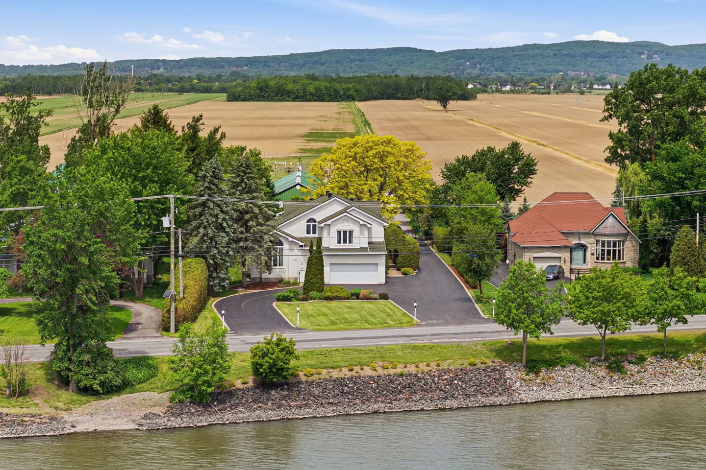 Aerial view of two houses along a river, with green trees, a paved driveway, and large agricultural fields in the background.