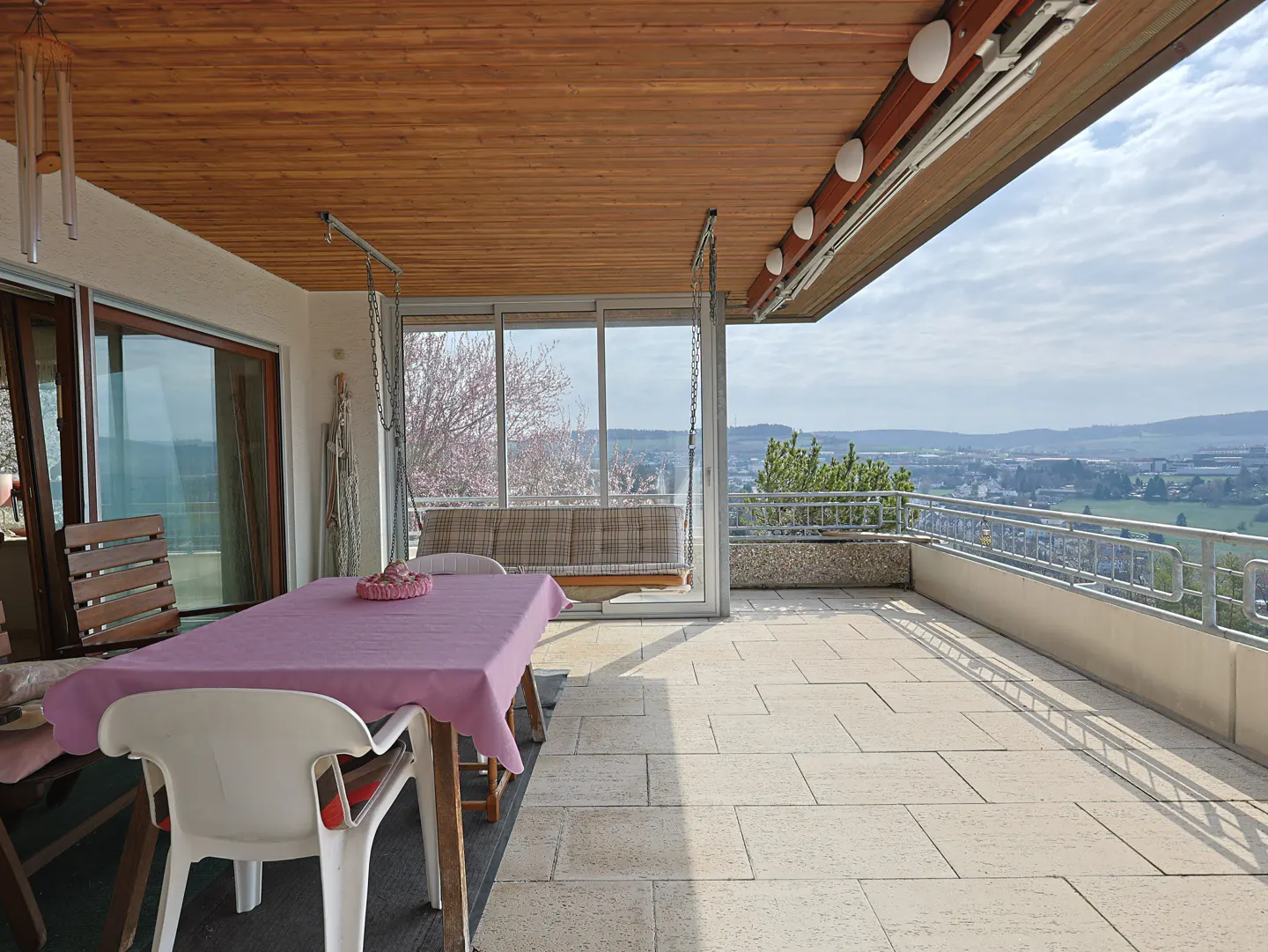 A covered patio with a table, chairs, and a swing overlooking a scenic view. The table is draped with a pink cloth.