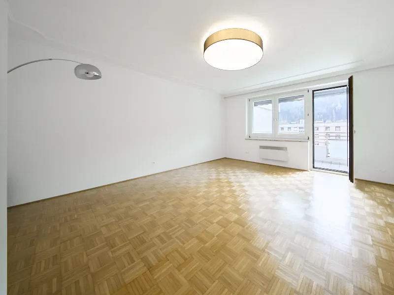 Bright, empty room with parquet floors, white walls, and a large window and balcony door letting in natural light. A modern lamp arches over the room.