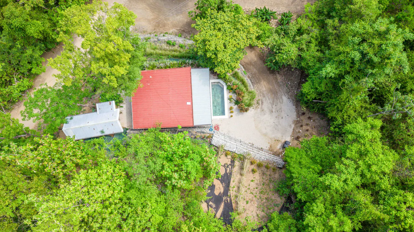 Aerial view of a house with a red roof and a pool, surrounded by lush green trees.