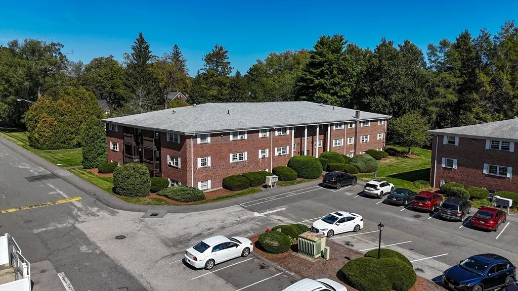 Aerial view of a two-story red brick apartment building with a gray roof and white trim, surrounded by green trees and a parking lot.