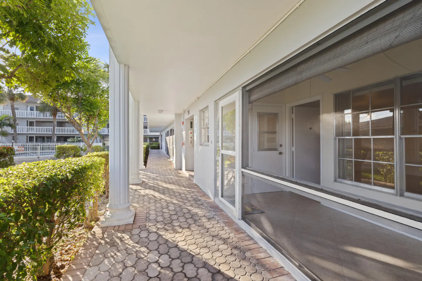 Exterior view of a white condo building with a brick walkway, columns, and green bushes. A screened-in porch is visible.