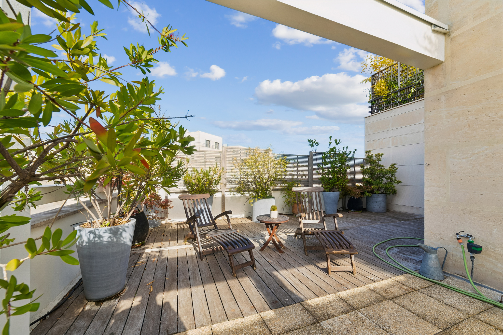 Sunny rooftop terrace with wooden decking, two lounge chairs, a small table, and various potted plants under a blue sky. [AI-generated text]