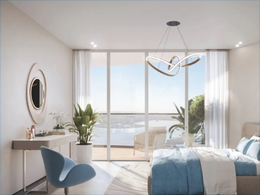 Bedroom with blue bedding, a vanity with a mirror, and a modern light fixture. Large windows offer a view of the ocean.