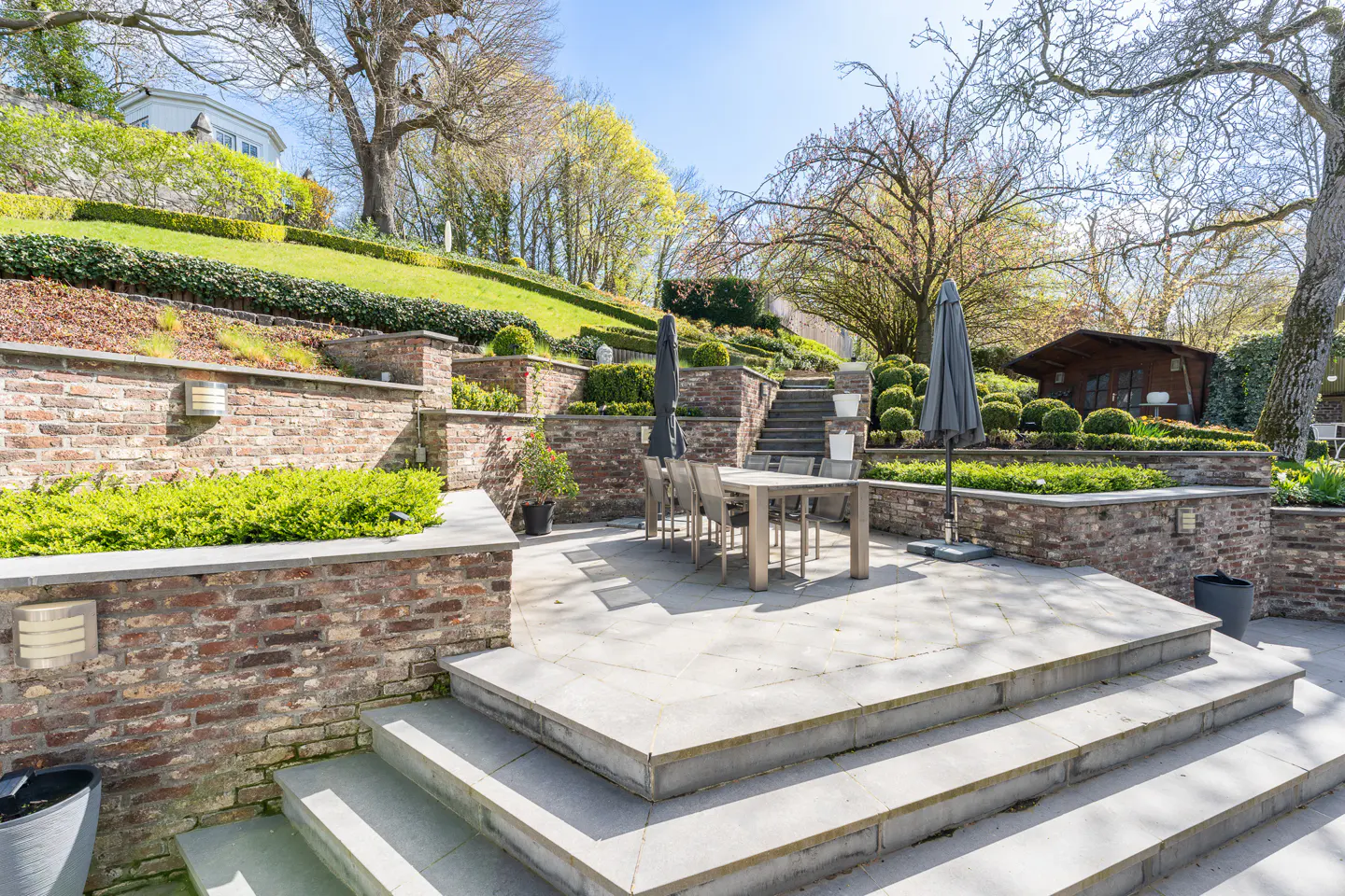 Outdoor patio with a table, chairs, and umbrellas on a multi-tiered stone terrace with brick walls and green landscaping.