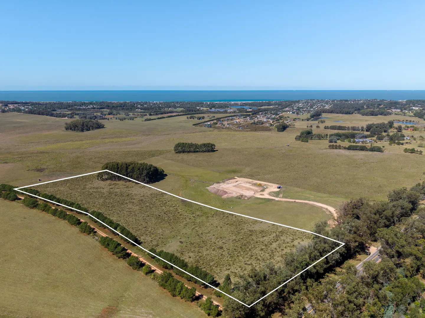 Aerial view of a large, grassy lot outlined in white, with trees, ocean, and blue sky in the background.