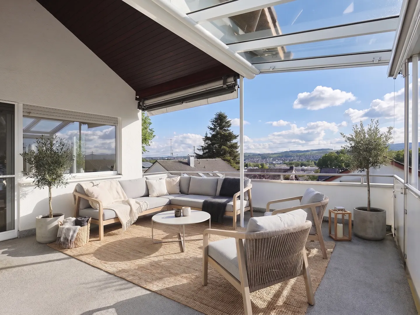Outdoor patio with a glass roof, featuring a gray sectional sofa, chairs, and a round table on a woven rug. A scenic view is visible.