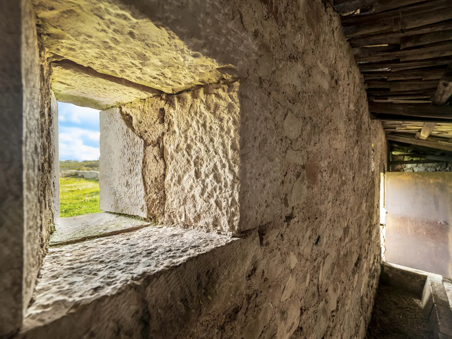 Stone wall with a window looking out onto a green field under a blue sky.