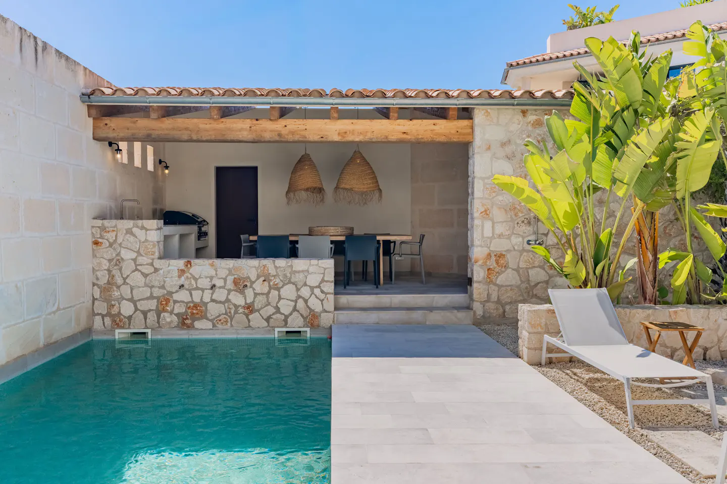 Outdoor pool area with stone walls, a covered dining space with straw pendant lights, and a white lounge chair under a clear blue sky.