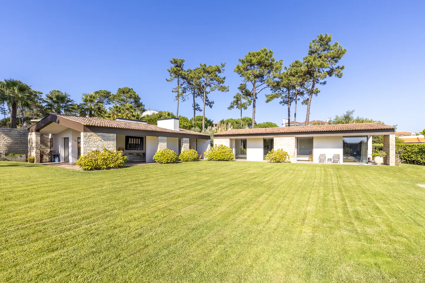 Two modern, single-story homes with white walls and brown tile roofs sit on a large green lawn under a clear blue sky.