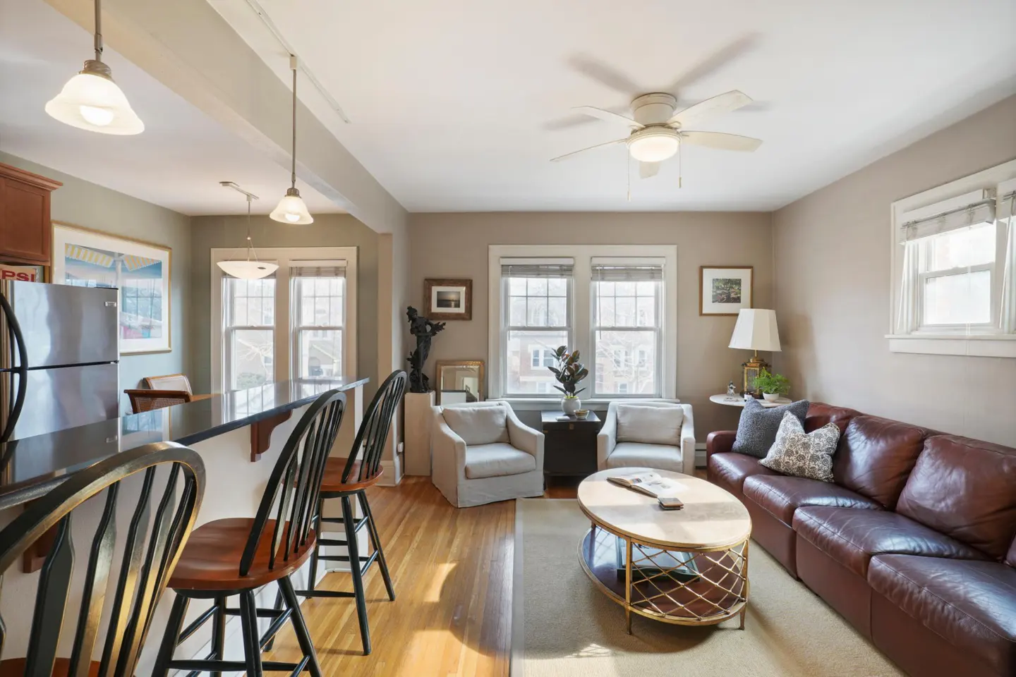 Open-concept living room with a brown leather sofa, two white armchairs, and a kitchen island with bar stools.