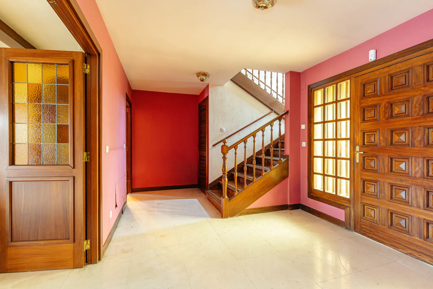 Interior view of a home's foyer with pink walls, a wooden staircase, and a decorative wooden front door with glass panels.