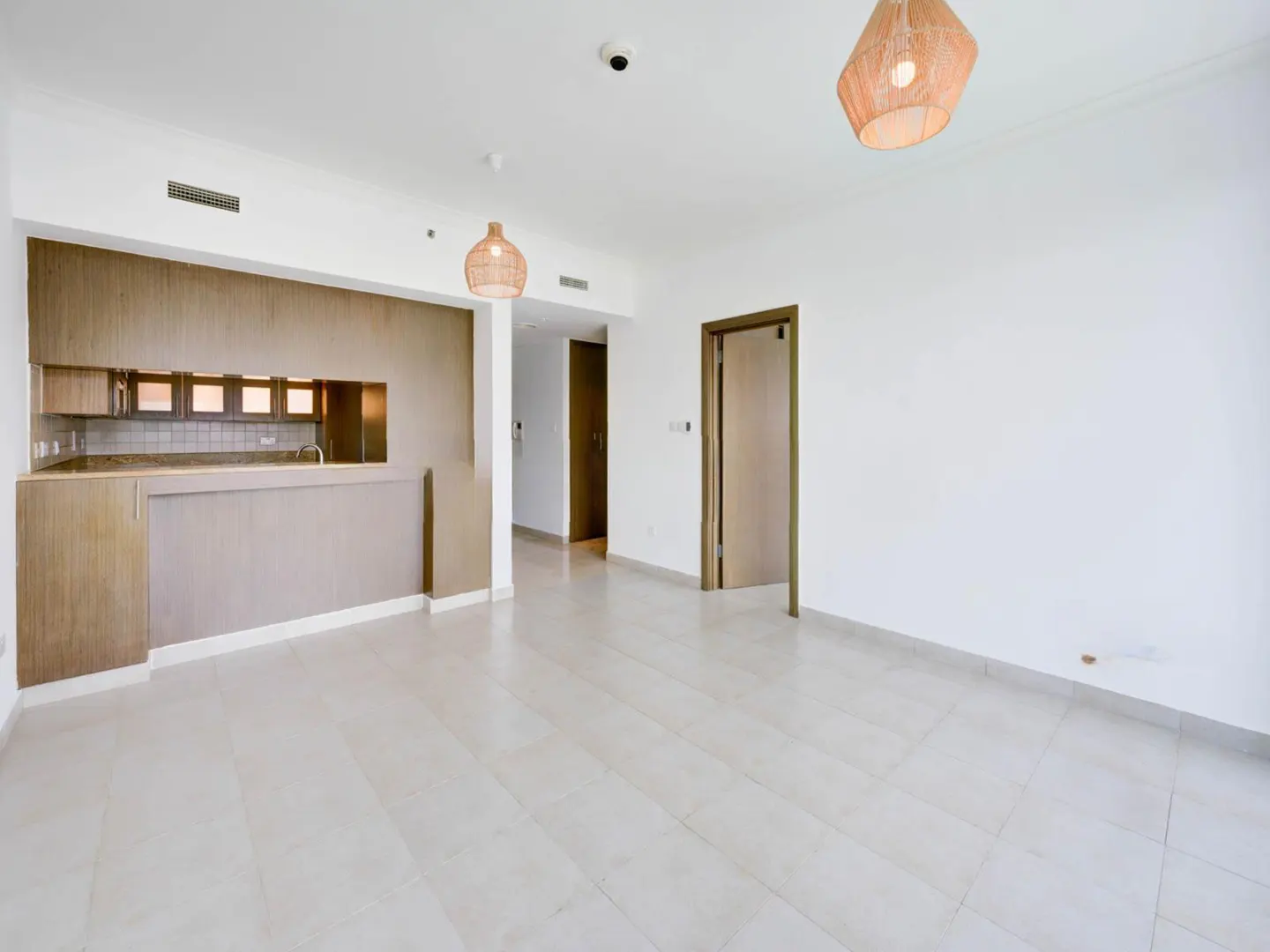 Bright, empty apartment with white walls, tile floors, and a kitchen area with wood cabinets. Two woven pendant lights hang from the ceiling.