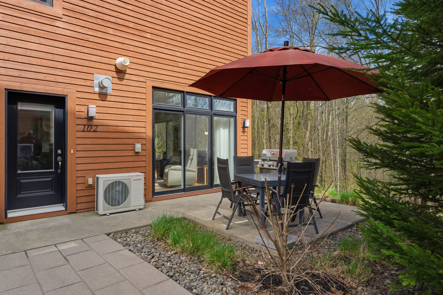 Exterior view of a brown house with a patio set, grill, and red umbrella. Door with number 102 is visible.