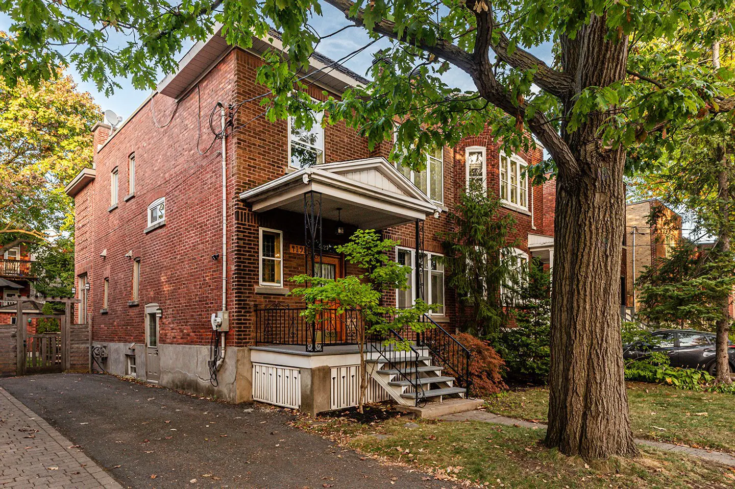 A two-story red brick house with a covered porch and black iron railings, surrounded by green trees and a paved driveway.