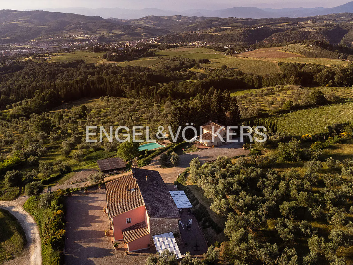 Aerial view of a Tuscan villa with a pool, surrounded by olive groves and vineyards. The villa has a red tile roof and a stone facade. Rolling hills in the background.