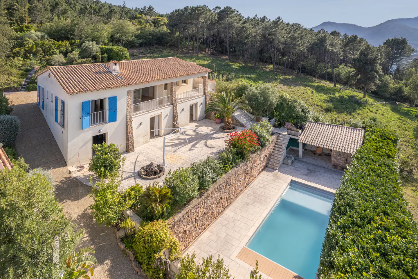 Aerial view of a two-story white house with blue shutters, a red tile roof, a pool, and lush greenery.