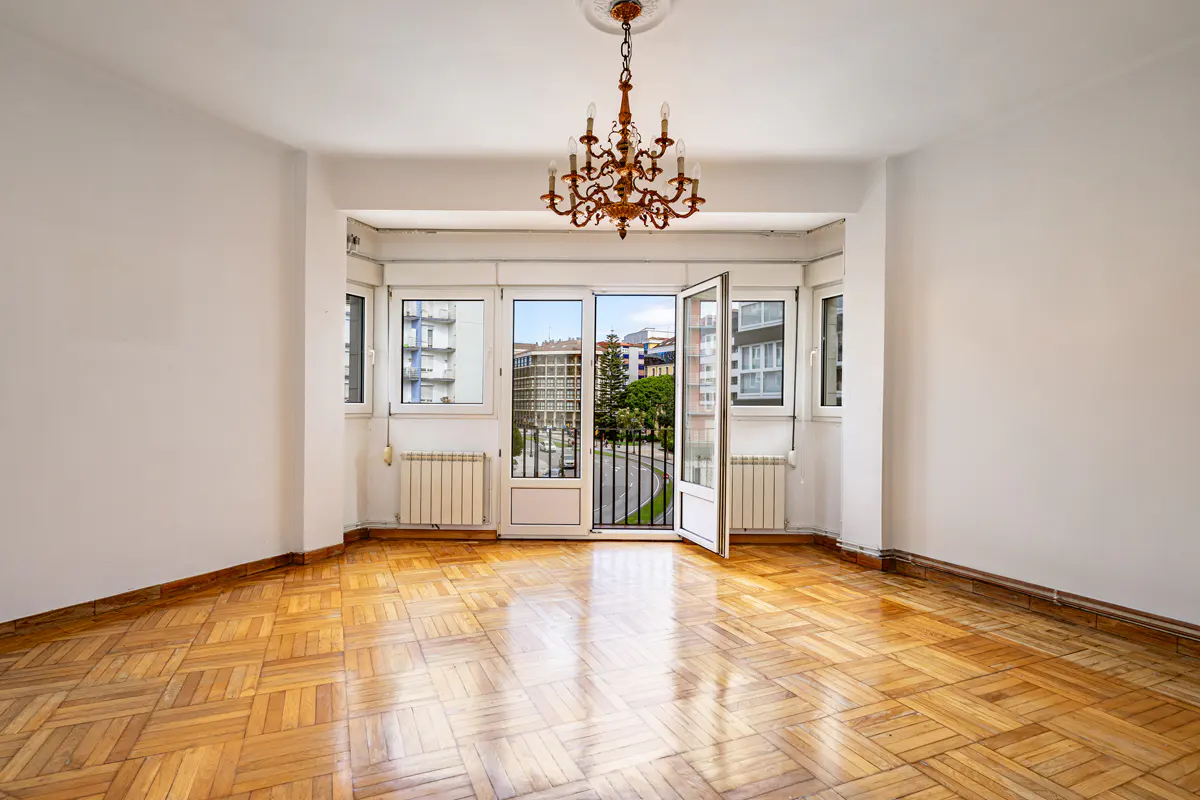 Empty room with parquet floor, white walls, and a balcony with an open door overlooking a city street. A chandelier hangs from the ceiling.