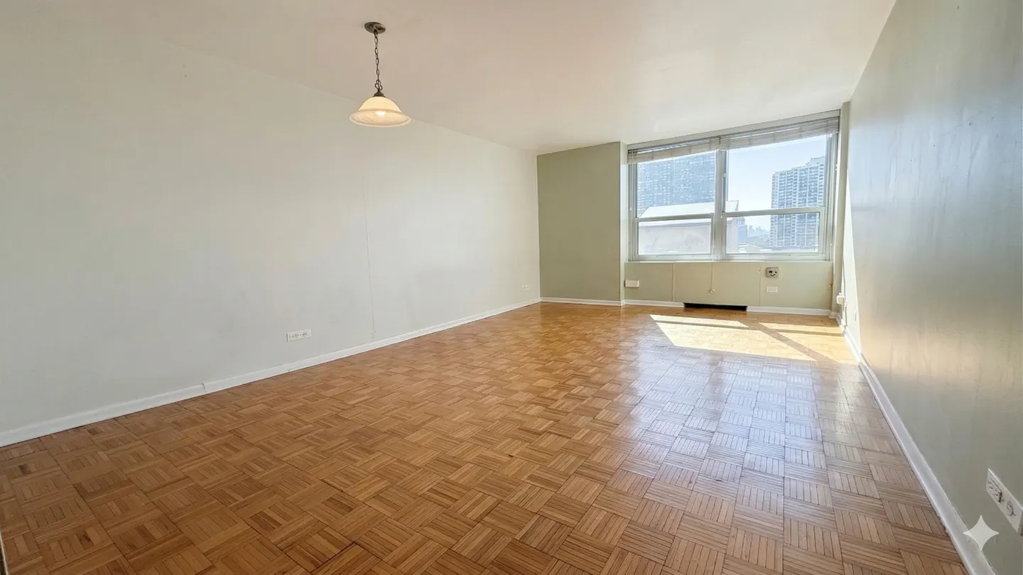 Bright, empty room with parquet floors, white walls, and a large window showing a city view. A pendant light hangs from the ceiling.