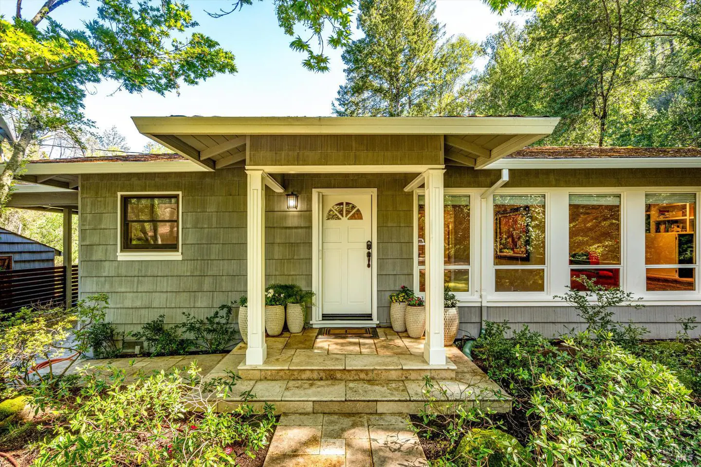 Front view of a house with gray siding, a white door, and a stone walkway. Green trees surround the house.