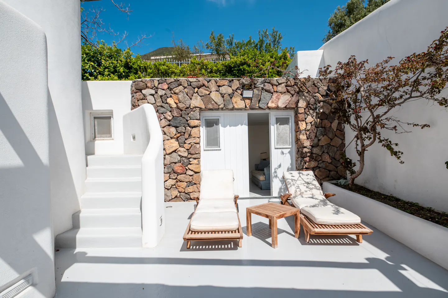 Outdoor patio with two lounge chairs, a small table, and a stone wall with a white door in Santorini, Greece.
