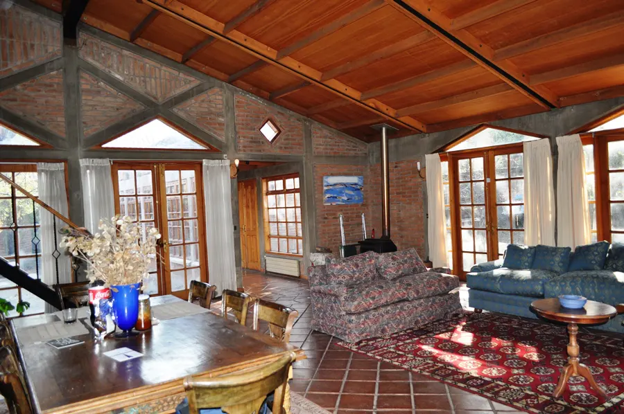 Open-plan living room with brick walls, wood ceiling, and tile floor. A dining table and sofas are visible. Natural light fills the space.