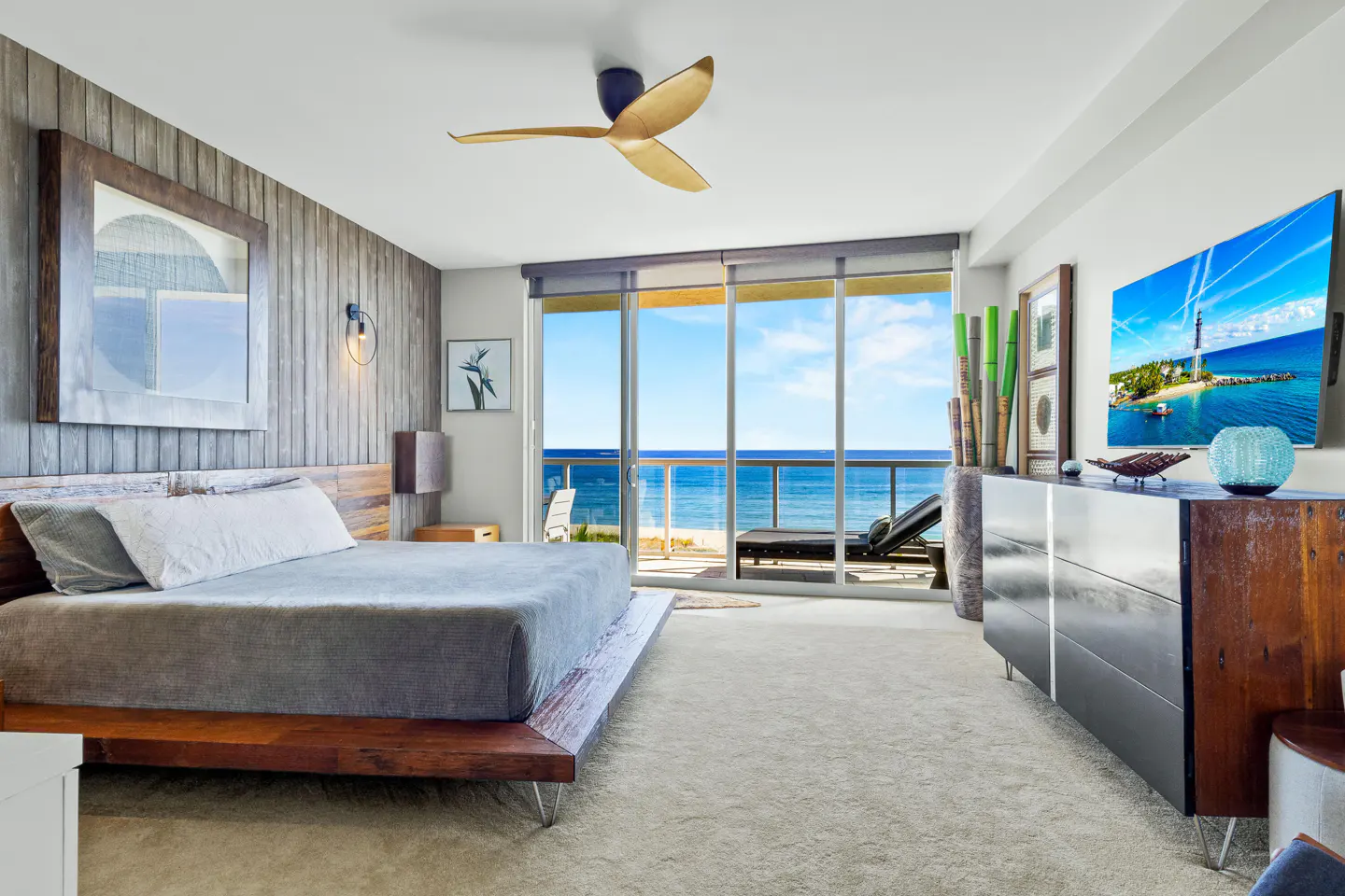 Bedroom with wood accents, a gray bed, and a ceiling fan. Sliding glass doors open to a balcony with ocean views. A TV is mounted on the wall.