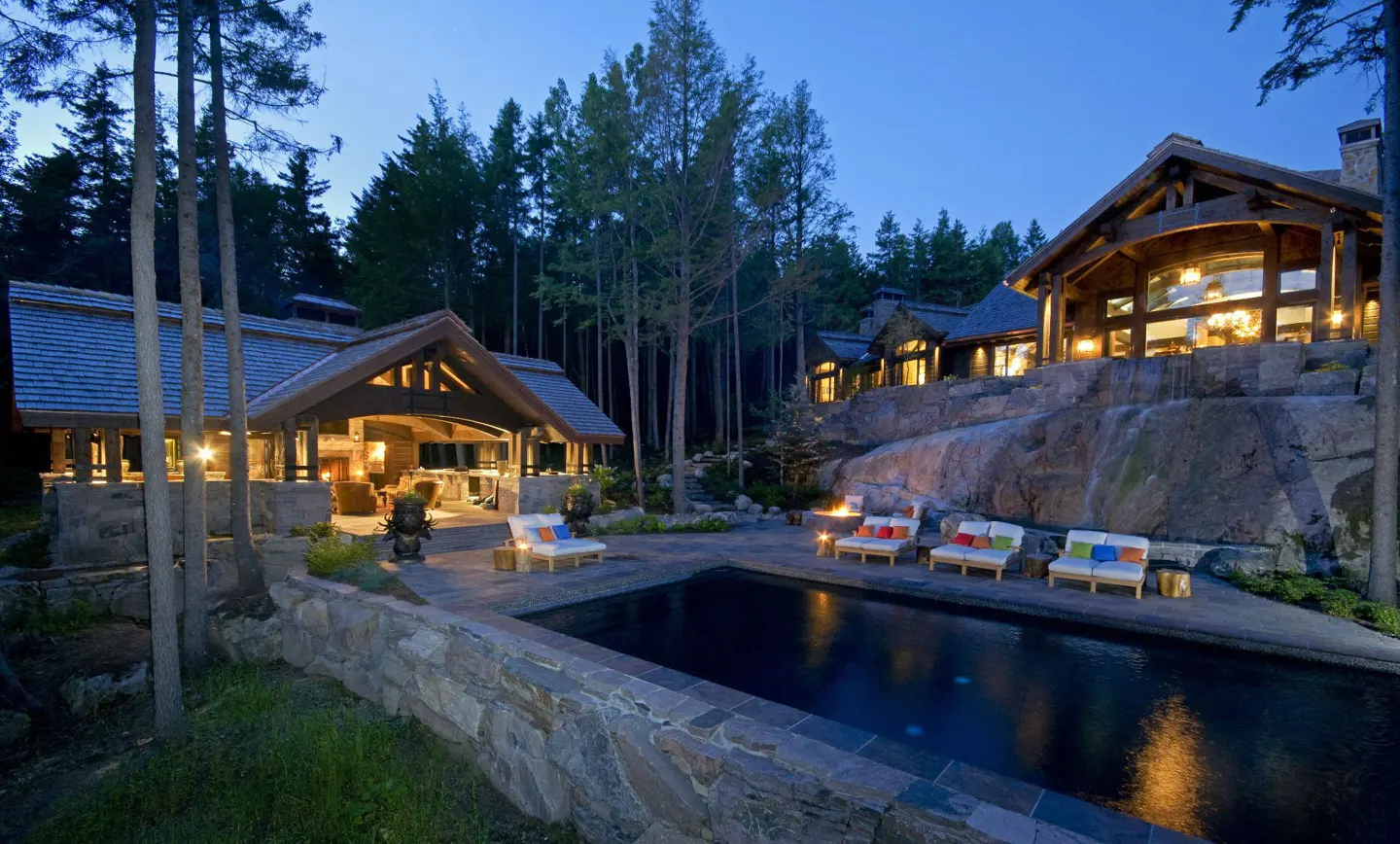 Luxury home exterior at dusk with a pool, lounge chairs, and an outdoor living area surrounded by trees.