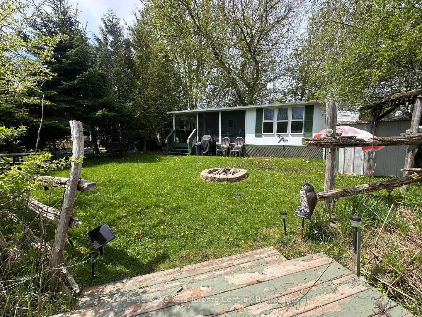 A white house with green shutters sits behind a green lawn with a fire pit. A wooden fence and owl statue are in the foreground.