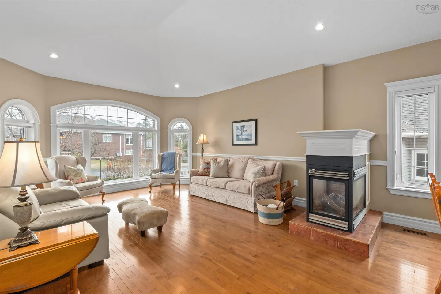 Bright living room with hardwood floors, beige walls, and white trim. Large windows offer outdoor views. Furnishings include sofas, chairs, and a fireplace.