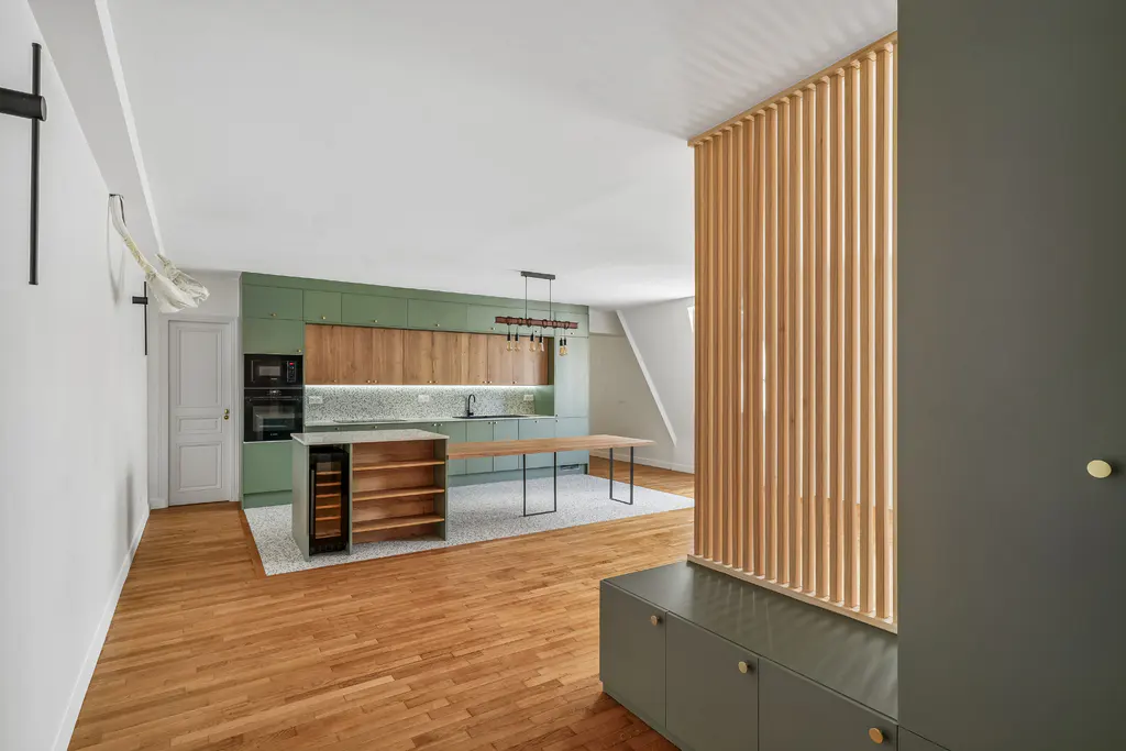 A modern kitchen with green cabinets, wood accents, and a light wood slat partition. A wood floor and island add warmth.