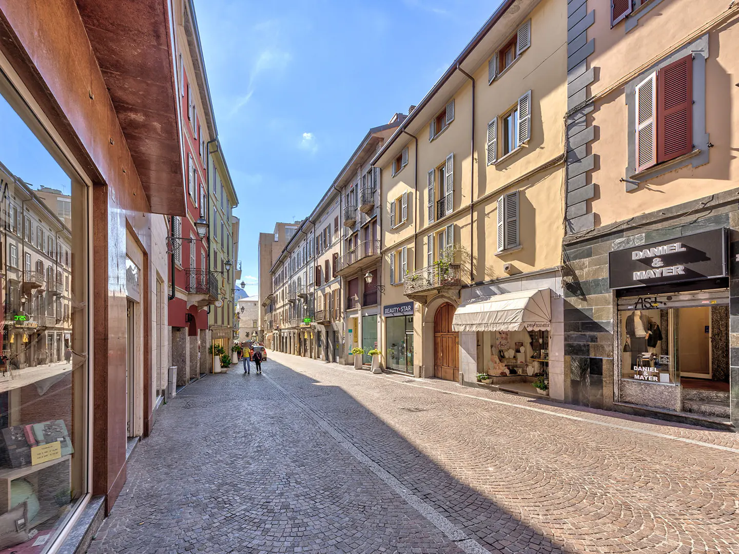 Cobblestone street lined with shops in Italy. Buildings are yellow, red, and green. Blue sky above.