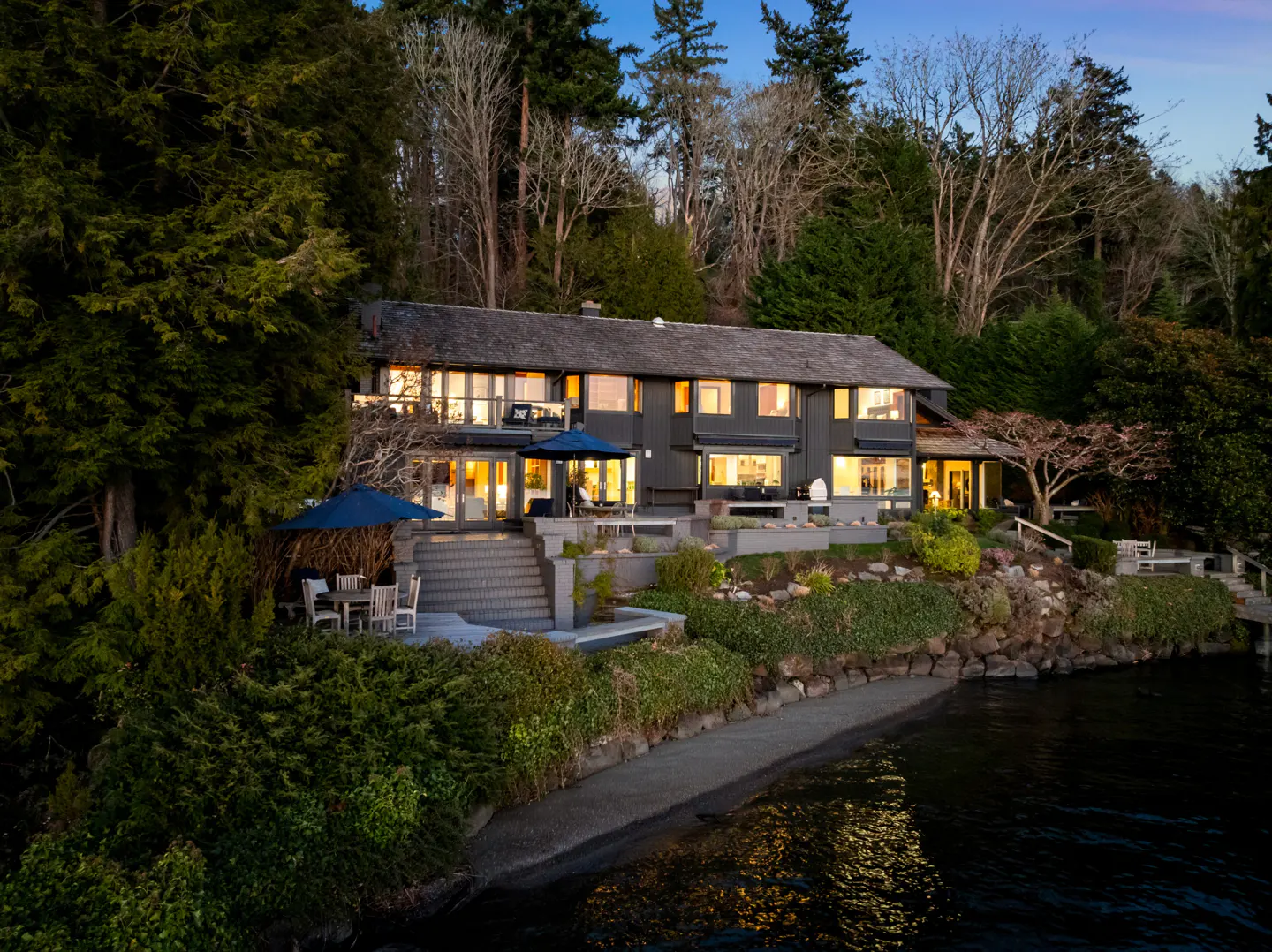 A two-story gray house with a shingled roof sits on a waterfront property with trees and a beach.