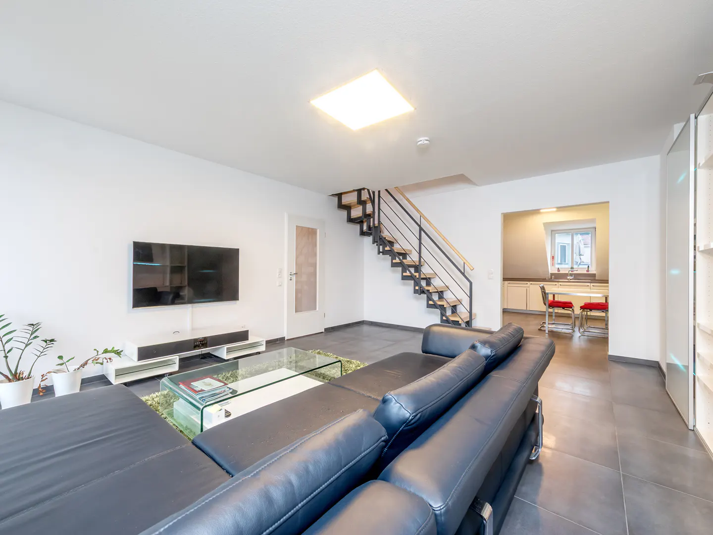 Bright living room with a black leather sofa, glass table, TV, and stairs leading to the upper floor. A kitchen is visible in the background.