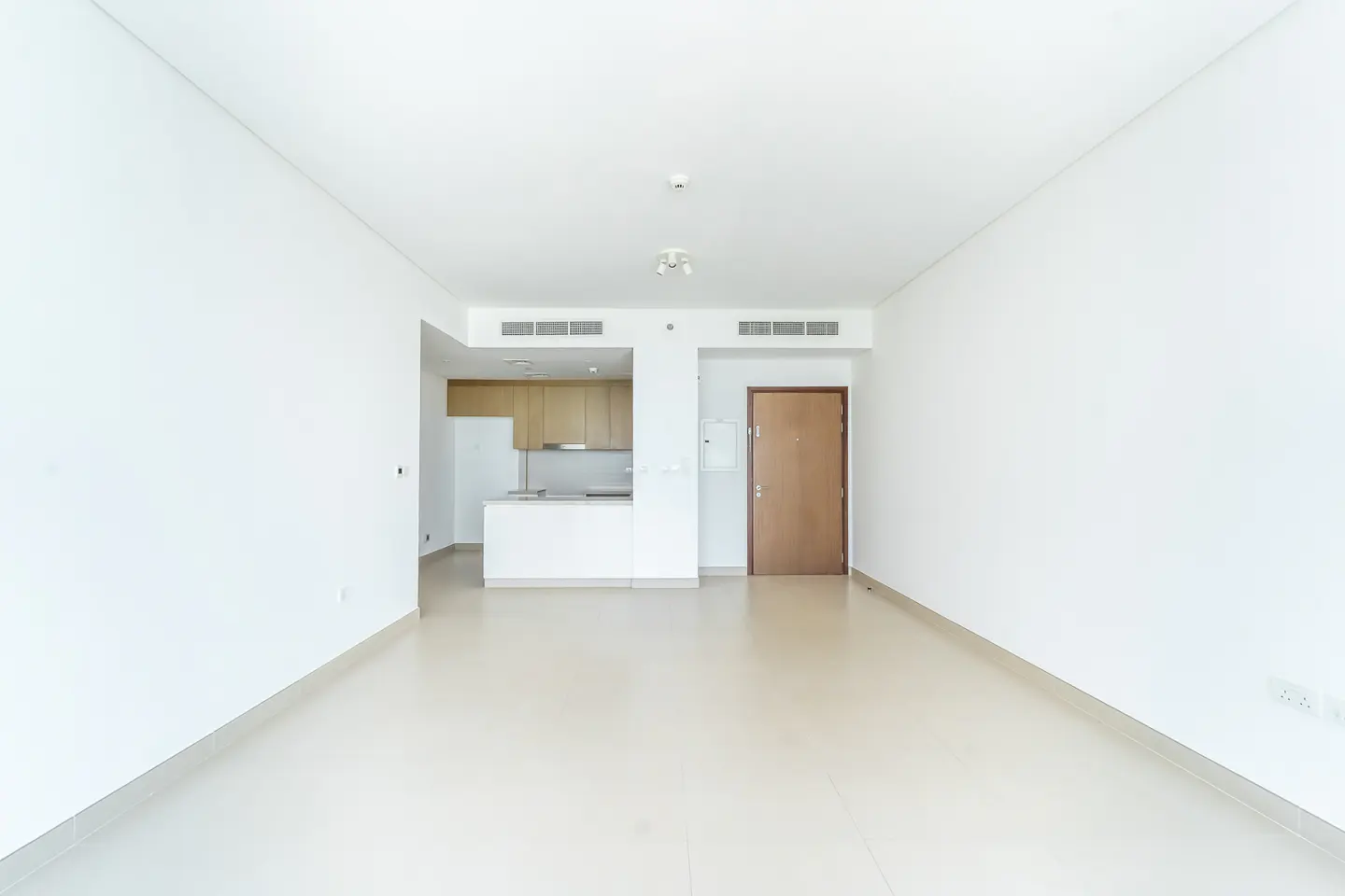 Empty apartment with white walls and tile floors. A kitchen with light wood cabinets is visible in the background. A brown door is on the right.