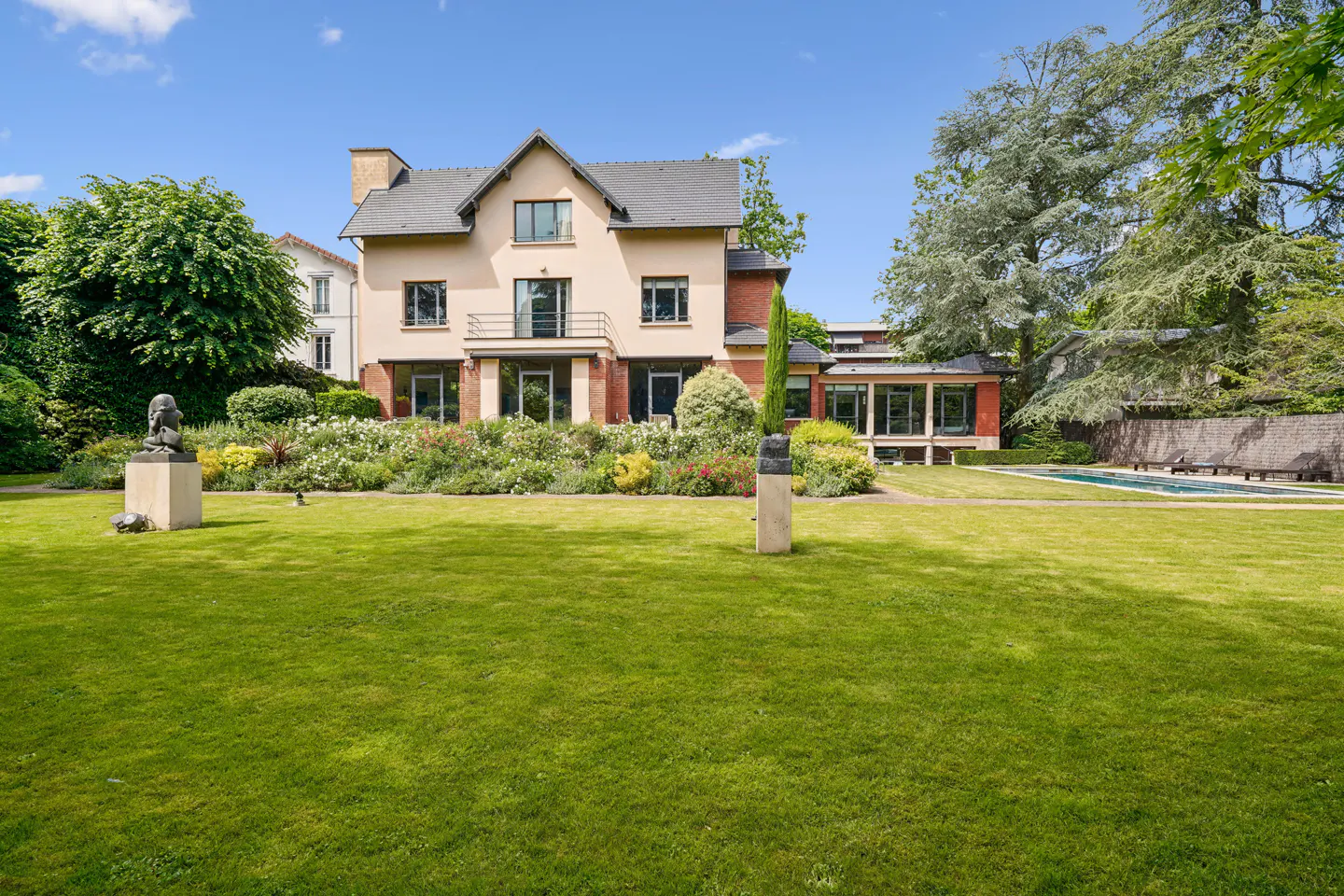 Large beige house with a dark roof, lush garden, sculptures, and a swimming pool on a sunny day.