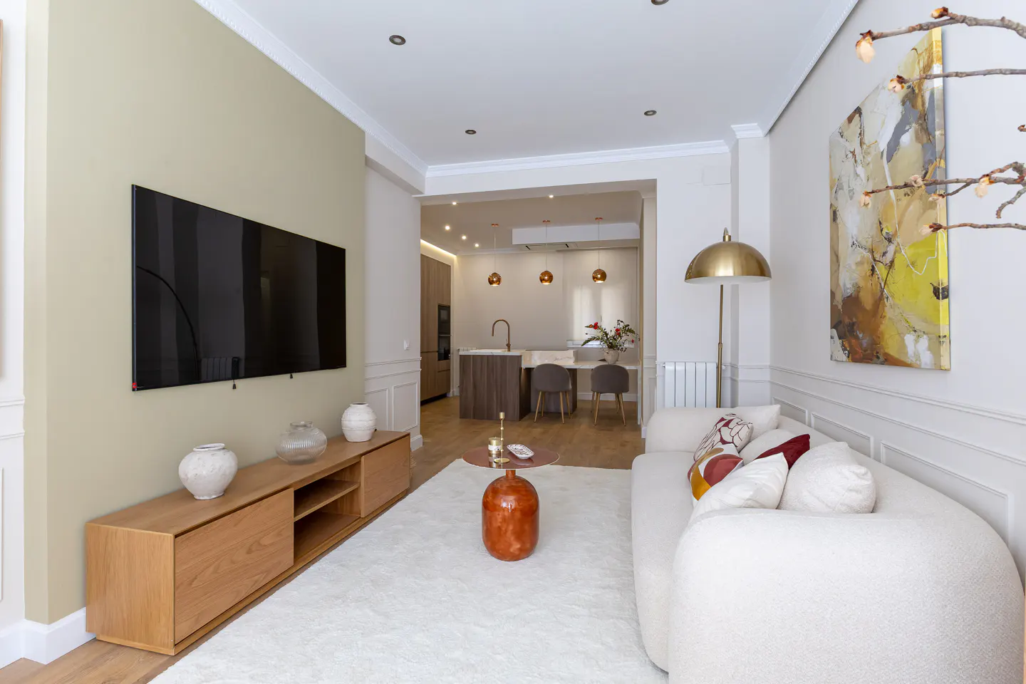 Bright, modern living room with a large TV, white sofa, and open kitchen in the background. A gold floor lamp stands near an abstract painting.