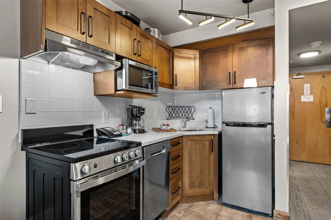 A compact kitchen with wood cabinets, stainless steel appliances, and white tile backsplash. A black stove and oven are on the left.