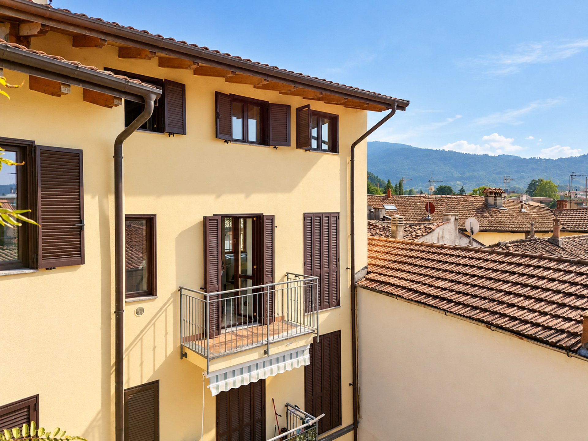 Yellow building exterior with brown shutters, a small metal balcony, and a view of tiled rooftops and distant mountains. [AI-generated text]
