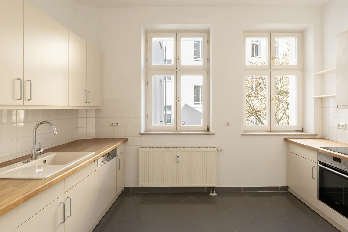Bright kitchen with white cabinets, wood countertops, and two windows. A sink and oven are visible. Gray tile floor.